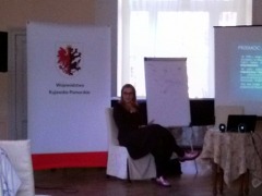 siedząca na krześle prowadząca szkolenie, pani Kinga Mickiewicz-Stopa, podczas przedstawiania prezentacji, obok widoczny roll-up województwa, fot. Biuro Wsparcia Rodziny i Przeciwdziałania Przemocy